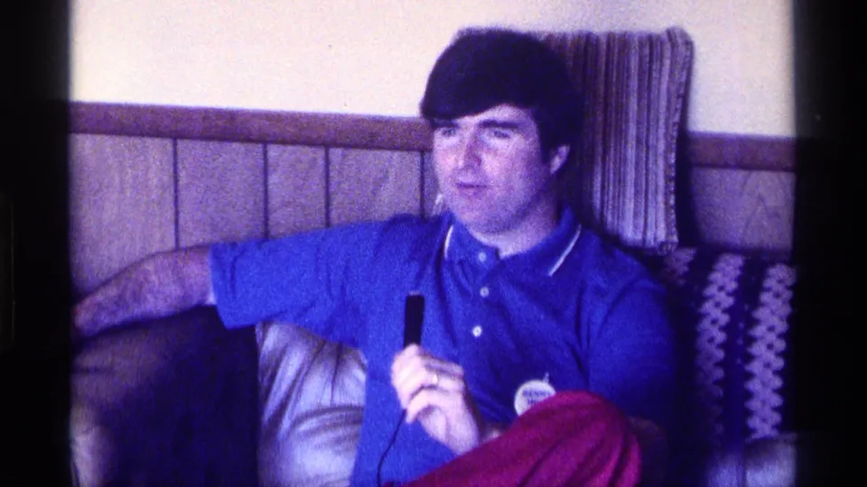 The seated man appears to listen or speak while holding a microphone in a 1970s Kentucky living room.