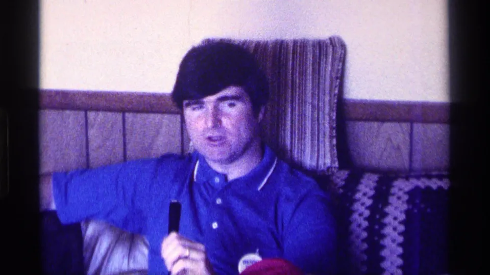 A man in a blue polo faces the camera from a couch in the Kentucky home-movie living-room scene.