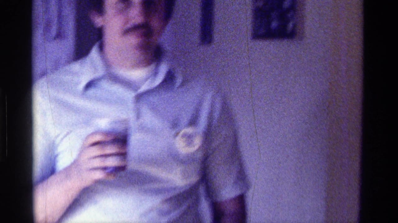 1976 Kentucky home-movie frame of a man in a light polo shirt holding a cup, with a button pinned to his shirt.