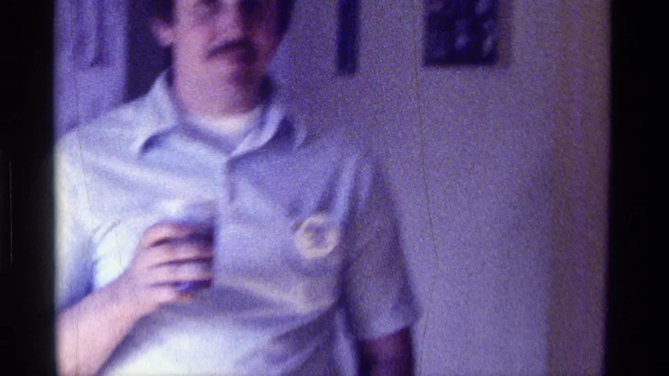 1976 Kentucky home-movie frame of a man in a light polo shirt holding a cup, with a button pinned to his shirt.