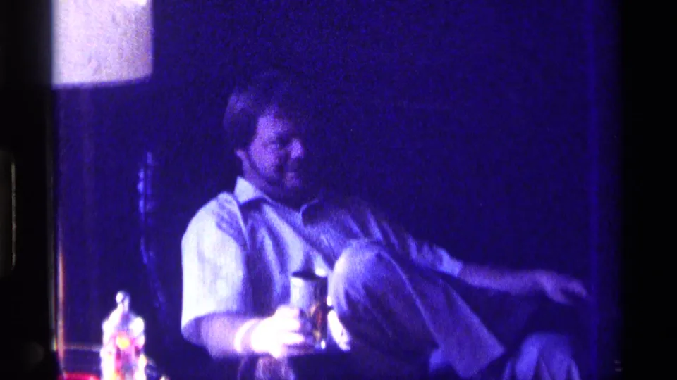 1976 Kentucky home-movie frame of a man seated in a dim interior, holding a drink while relaxing with one leg raised.
