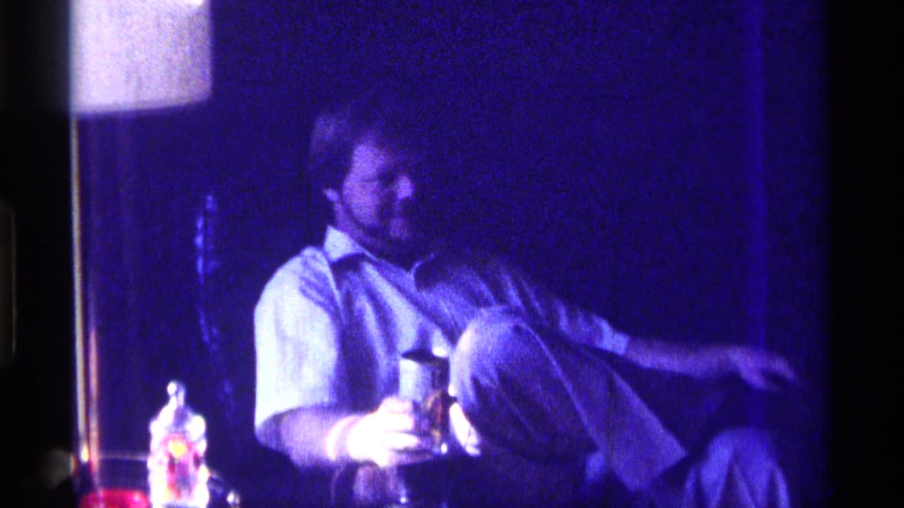 White man seated indoors in dim blue light, holding a drink near his lap.