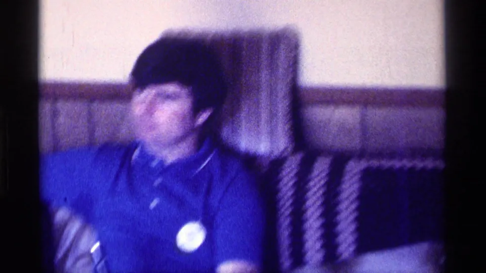 1976 Kentucky home-movie frame of a younger white man in a blue polo seated on a couch against wood paneling.