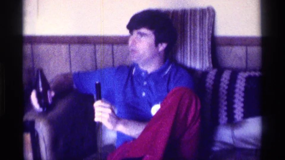 1976 Kentucky 8mm frame of a younger white man in a blue polo and red pants seated on a couch, holding a bottle while looking aside.