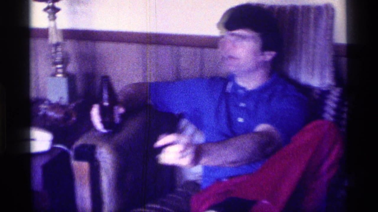 Man in a blue shirt and red pants sits on a couch holding a drink in a paneled living room.