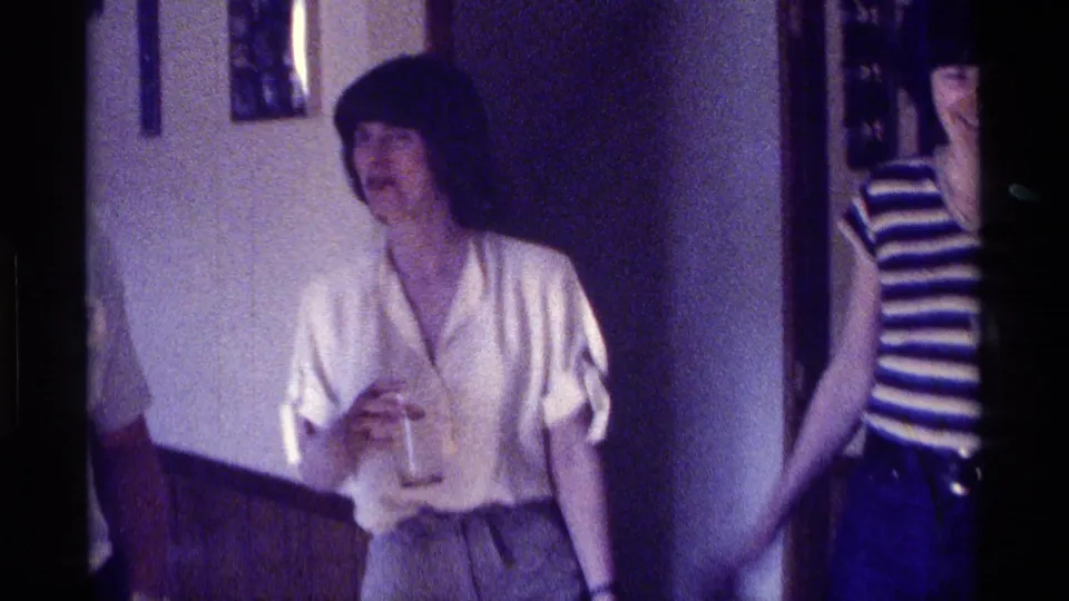 1976 Kentucky home-movie frame of a woman with a drink near a doorway, with another woman partly visible at the edge.