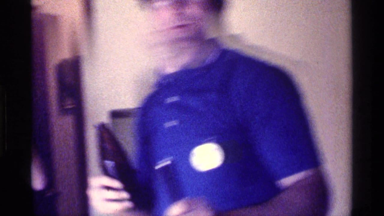 Blurred man in a blue polo shirt points toward a round button on his chest indoors.