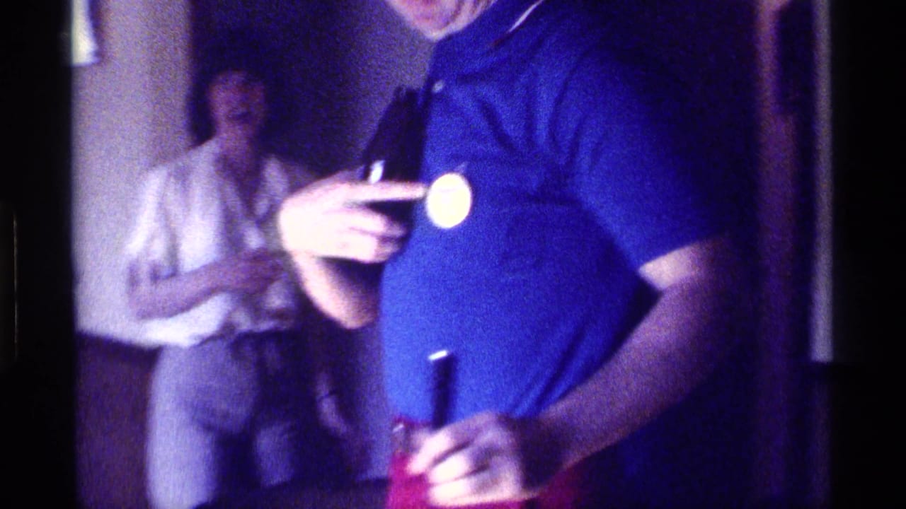 1976 Kentucky indoor party frame with a man showing a chest button and another adult standing behind him holding a cup.