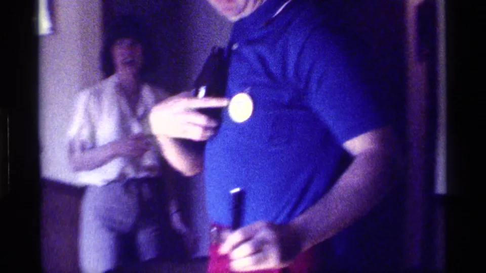 1976 Kentucky indoor party frame with a man showing a chest button and another adult standing behind him holding a cup.