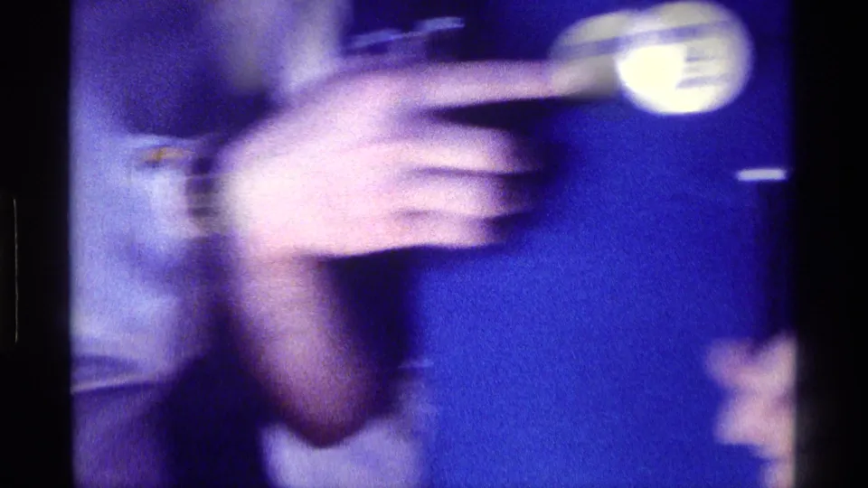Close, motion-blurred 1976 Kentucky home-movie frame focused on a hand pointing to a round button on a blue polo shirt.