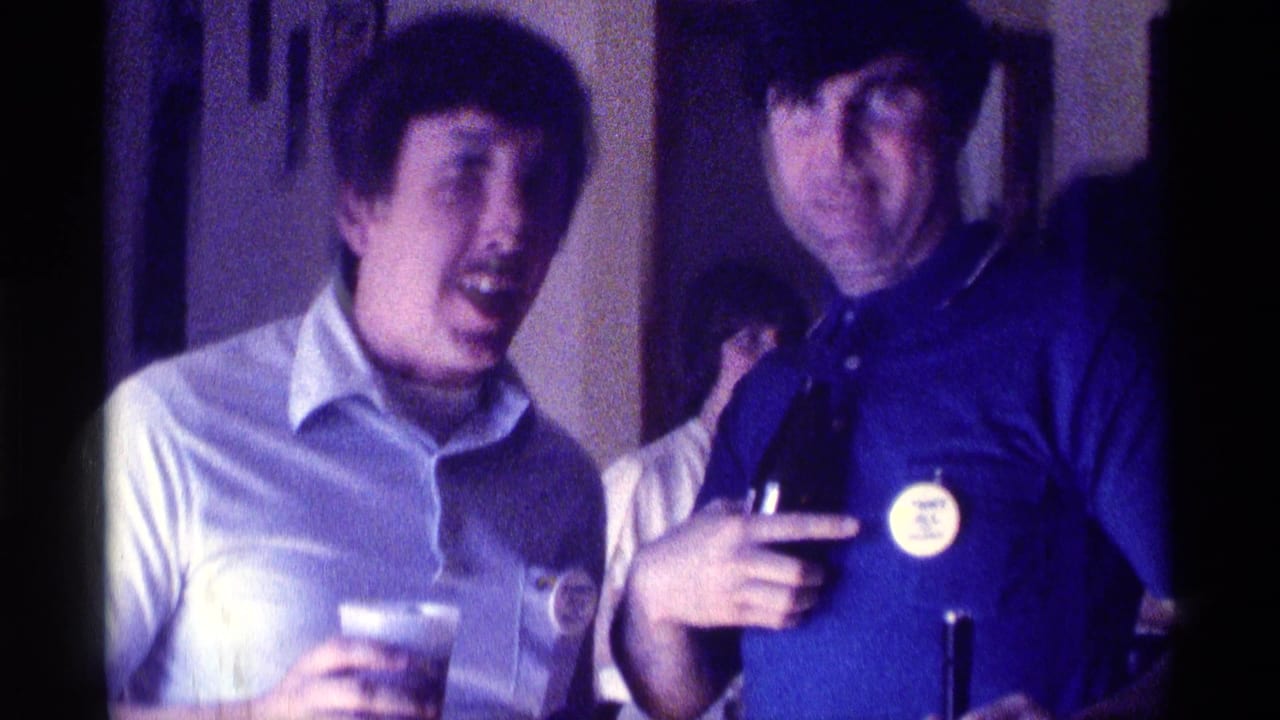 Two men indoors at a party, one holding a plastic cup and the other holding a bottle.