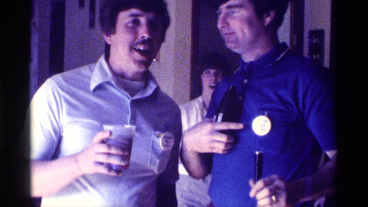 1976 Kentucky frame of two men in polo shirts at an indoor gathering, with a smiling woman behind them.