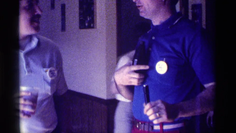 1976 Kentucky home-movie frame with cropped adult men socializing indoors, including drinks and a visible name badge.