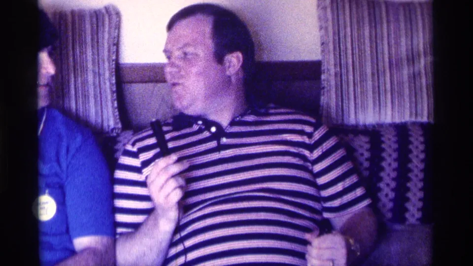 1976 Kentucky indoor party frame showing an adult man in a striped shirt holding a microphone near patterned curtains.