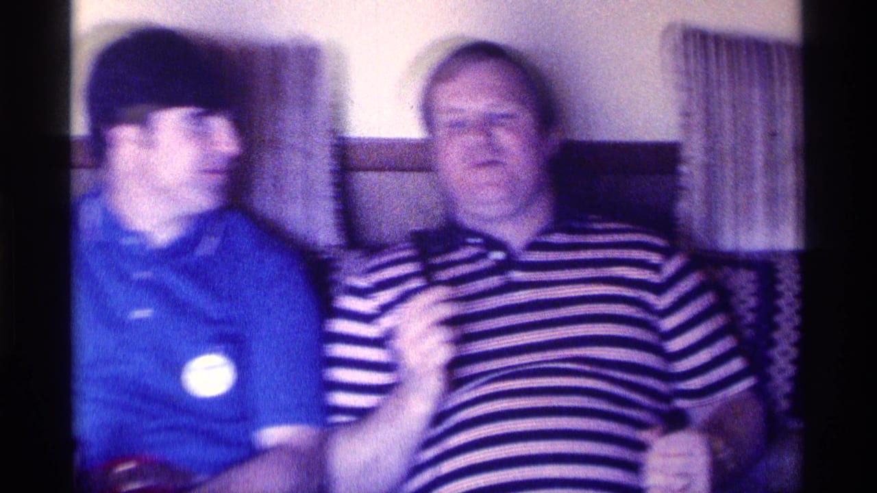 Two men seated on a couch, one in a blue polo and one in a striped polo holding a microphone.