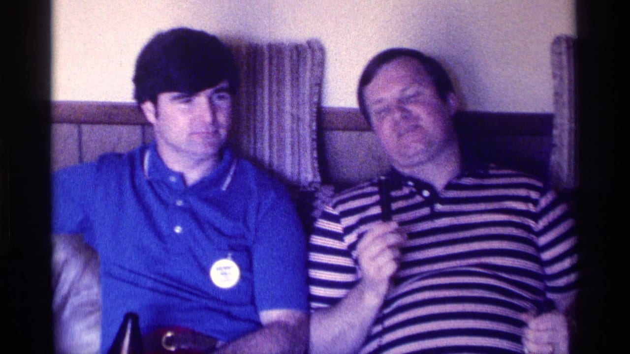 The striped-shirted man appears to speak into a handheld microphone beside a younger man in a blue polo in a Kentucky living room.
