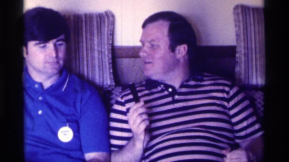 The older man in a striped polo turns toward the man in blue, holding a microphone during an informal 1976 indoor exchange.