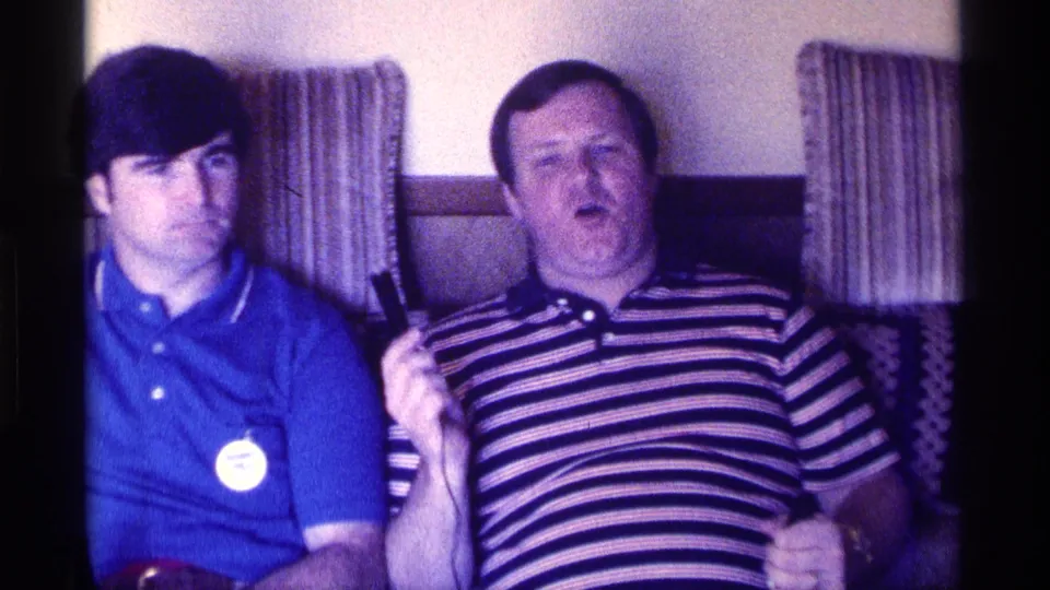 The striped-shirted man gestures with a handheld microphone beside the man in blue in a softly focused Kentucky home-movie frame.