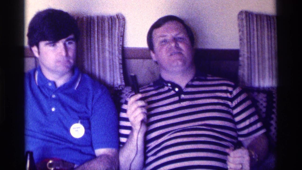 In a 1976 Kentucky home interior, two adult men sit closely on a couch, with the man in a striped polo holding a small microphone.