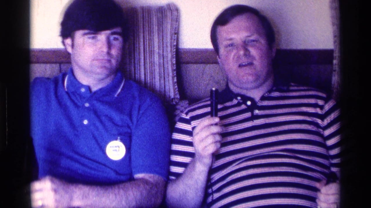 Man in striped shirt raises a small microphone beside a man in a blue shirt.