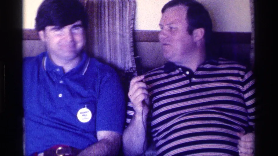 In the private living room setting, the man with the microphone turns toward the man in the blue polo as they sit on the couch.