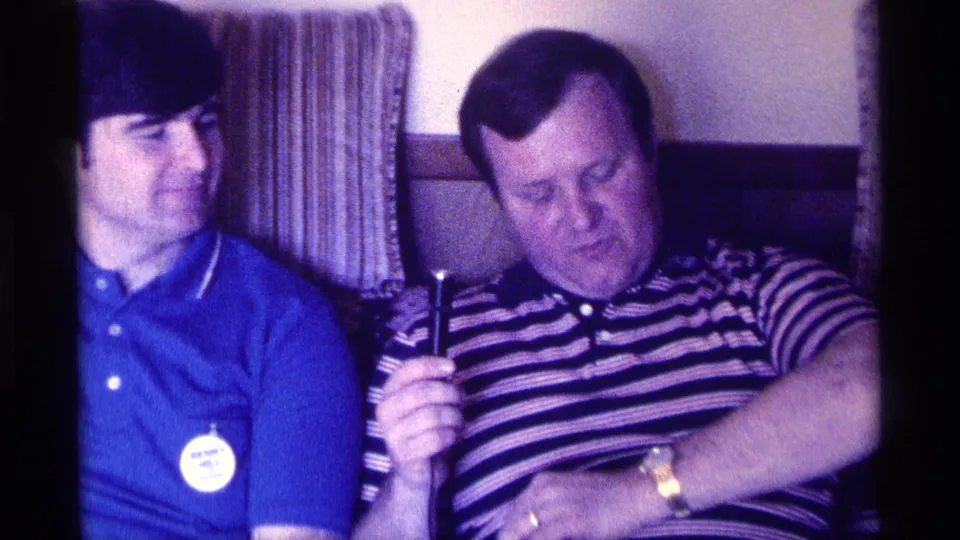 The man in the striped polo checks his wristwatch while holding the microphone, seated beside the man in the blue shirt in a 1976 Kentucky interior.