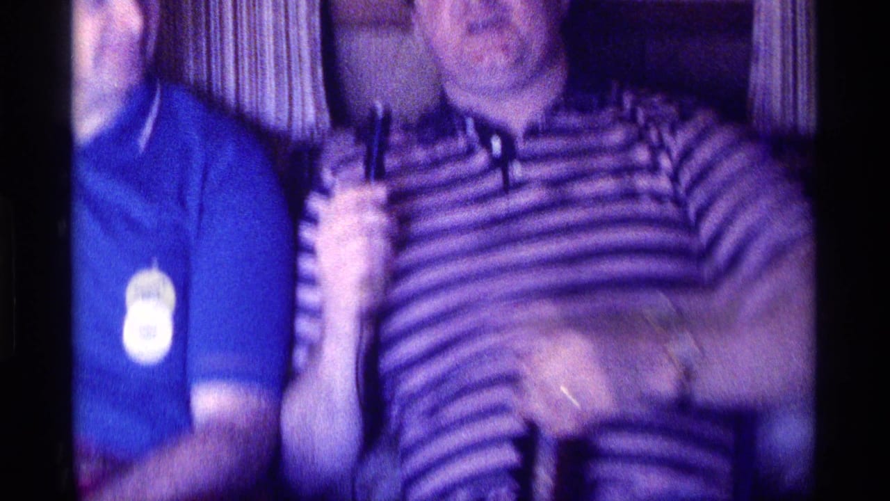 Kentucky 1976 8mm frame of two adult men seated indoors, with a striped polo shirt, blue shirt, curtains, and motion blur.