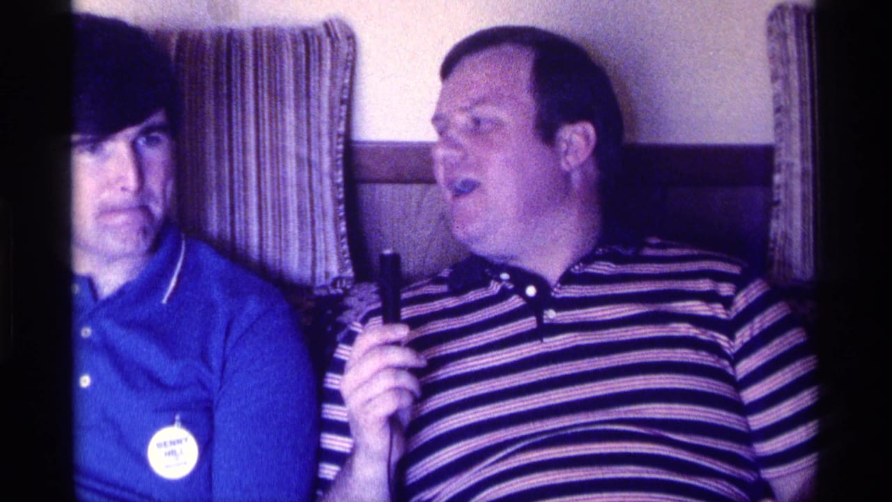 Two men sit side by side indoors as the man in stripes holds a small object and speaks.