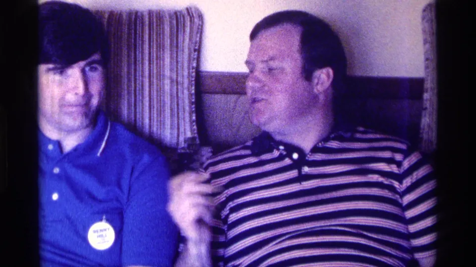 Kentucky 1976 8mm frame of two adult men seated on a couch during an indoor social gathering, with curtains behind them.