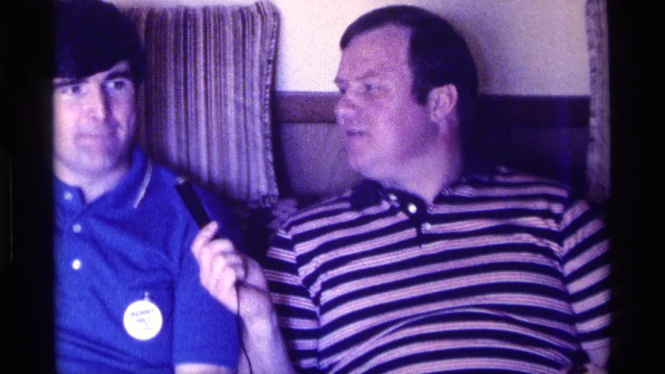 Kentucky 1976 home-movie frame of two adult men seated indoors, with the striped-shirted man holding up a small object.