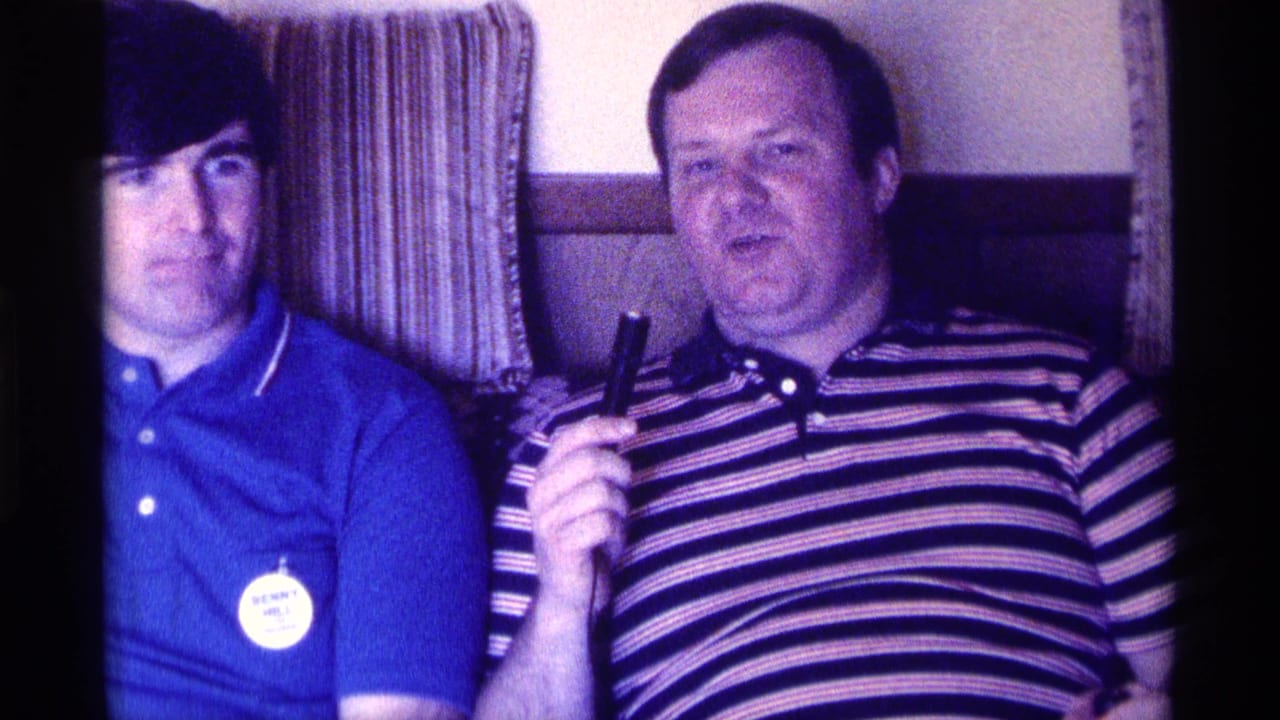 Two men sit on a couch while the man in a striped shirt holds a microphone.