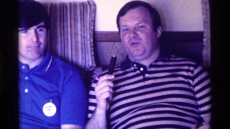 In 1976 Kentucky home-movie footage, a young man in a blue polo sits beside an older man in a striped polo holding a microphone.