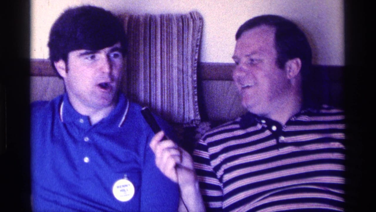 The interviewer in a blue polo appears to speak while the older man in a striped polo smiles beside him in a private interior.