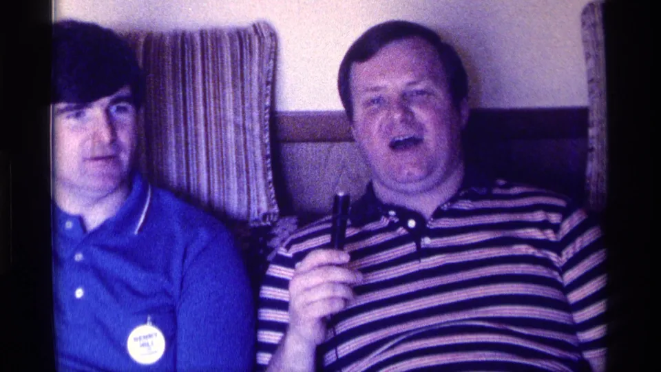 A close 8mm frame shows two men seated on a couch, with the striped-shirt man holding a microphone during the informal interview.