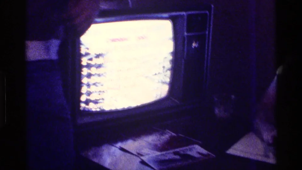 In a dim 1976 Kentucky home interior, a boxy television glows while dark furnishings and papers or objects sit below the screen.