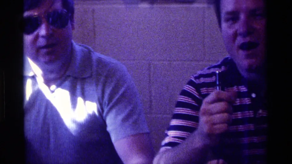Two adult men sit close together in a private Kentucky interior during the 1976 home-movie clip, one in sunglasses and a light shirt and the other in a striped polo shirt.