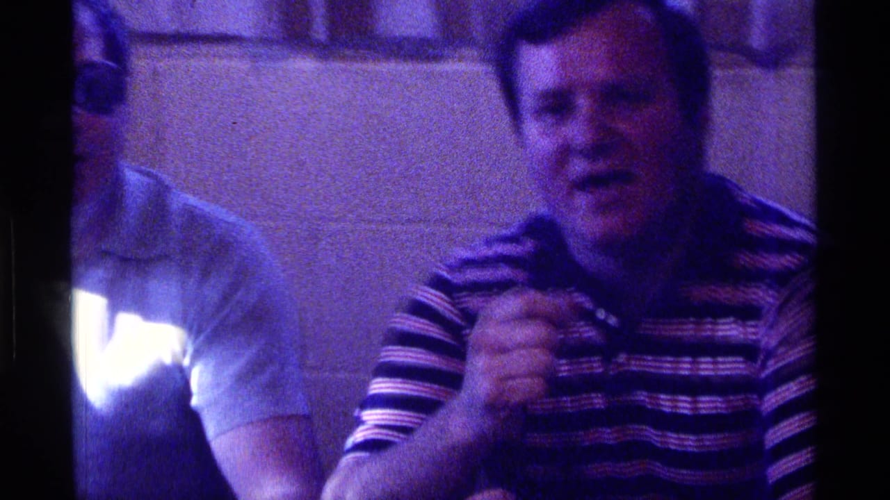 Two adult men seated indoors, one partly cropped and one holding a microphone.