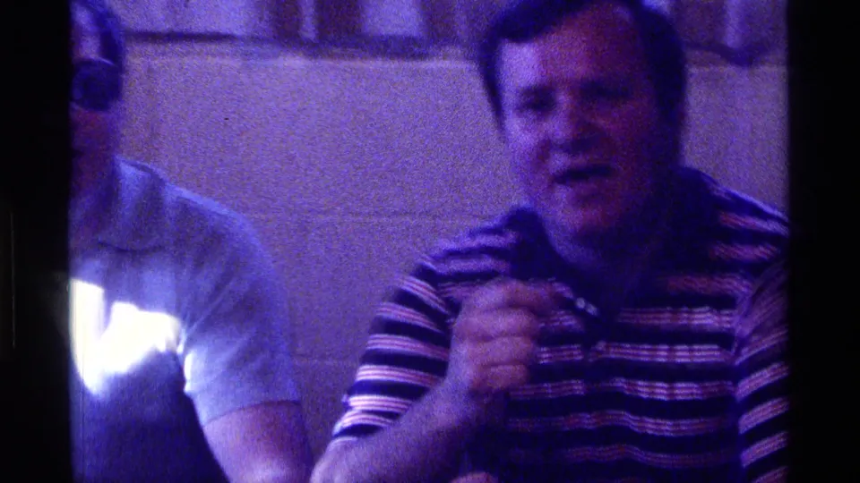 In 1976 Kentucky home-movie footage, a man in a striped polo holds a microphone beside another partly visible seated man indoors.