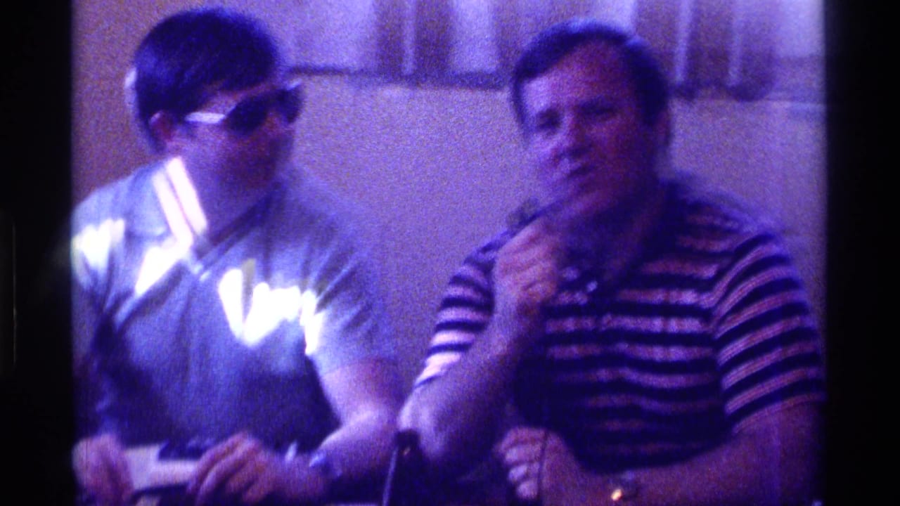 Two men sit indoors in 1976 Kentucky footage, with one wearing sunglasses and the other holding a microphone near his face.
