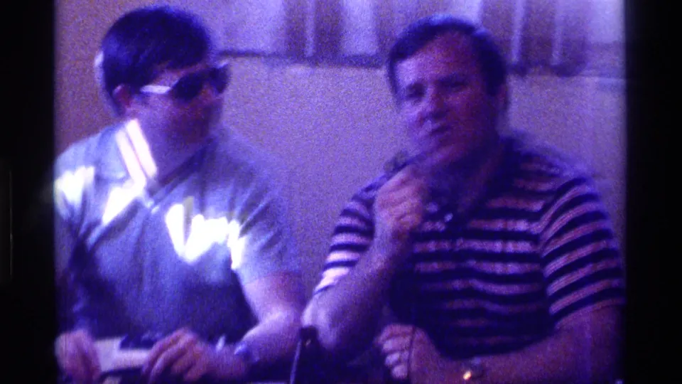 Two men sit indoors in 1976 Kentucky footage, with one wearing sunglasses and the other holding a microphone near his face.