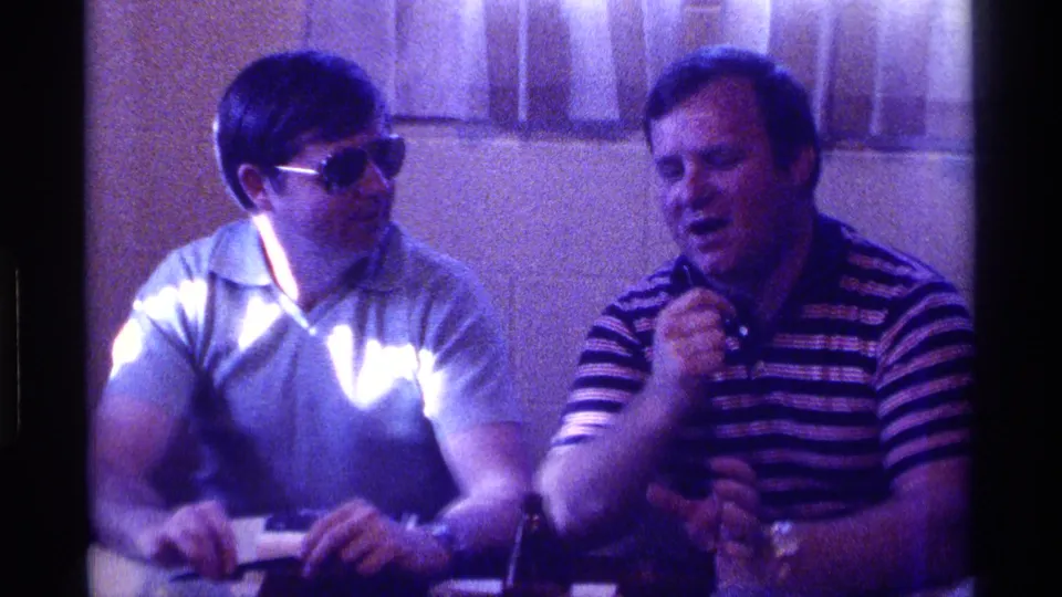 A 1976 Kentucky indoor home-movie frame shows a man in sunglasses seated beside a striped-shirt man holding a microphone.