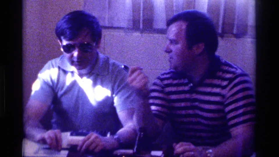 The striped-shirt interviewer turns toward the sunglasses-wearing man in this 1976 Kentucky indoor home-movie frame.