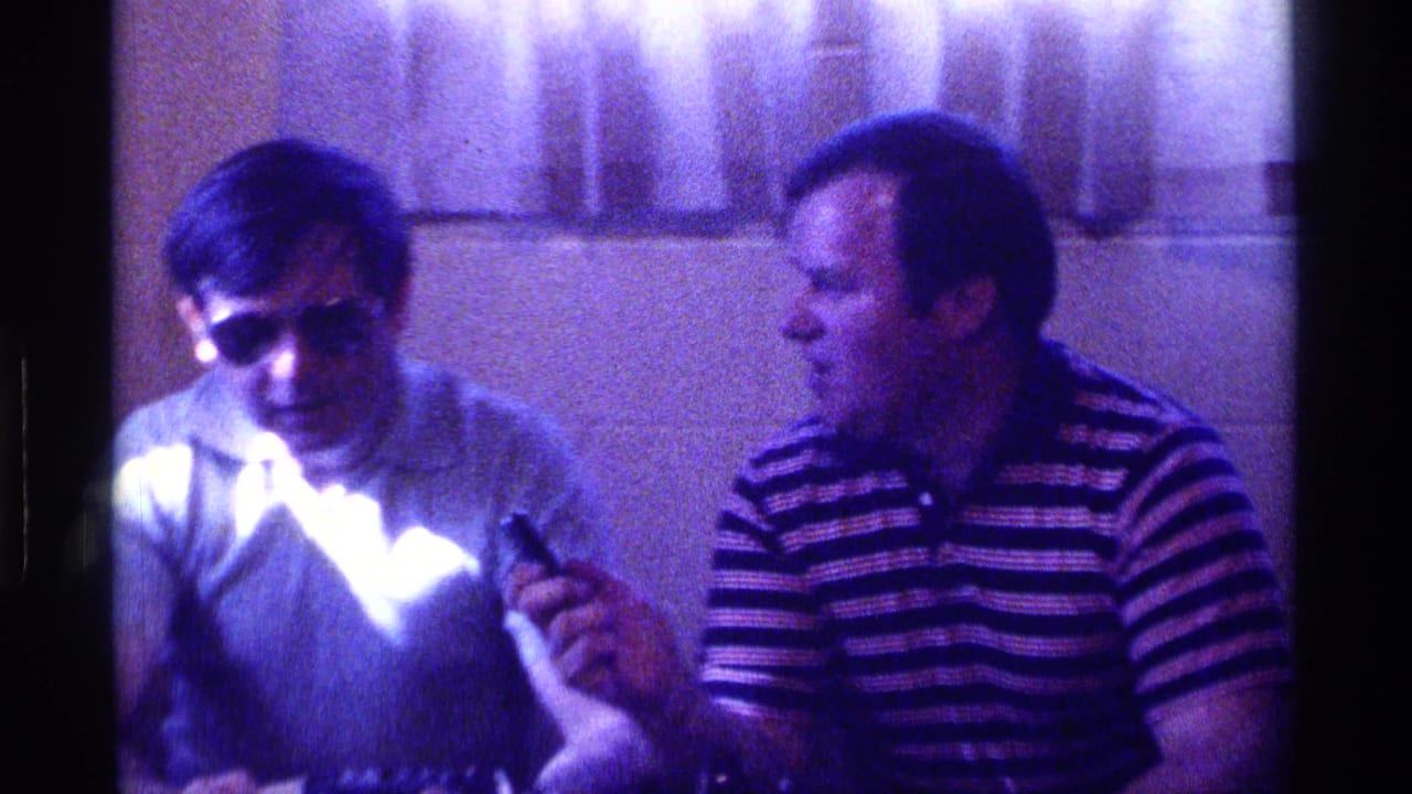 Two men sit indoors at a table as one holds a small microphone near the other.
