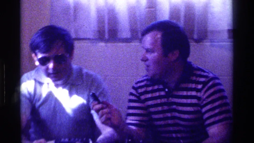 In 1976 Kentucky home-movie footage, two adult men sit in a dim room while the man in a striped polo holds a small microphone.