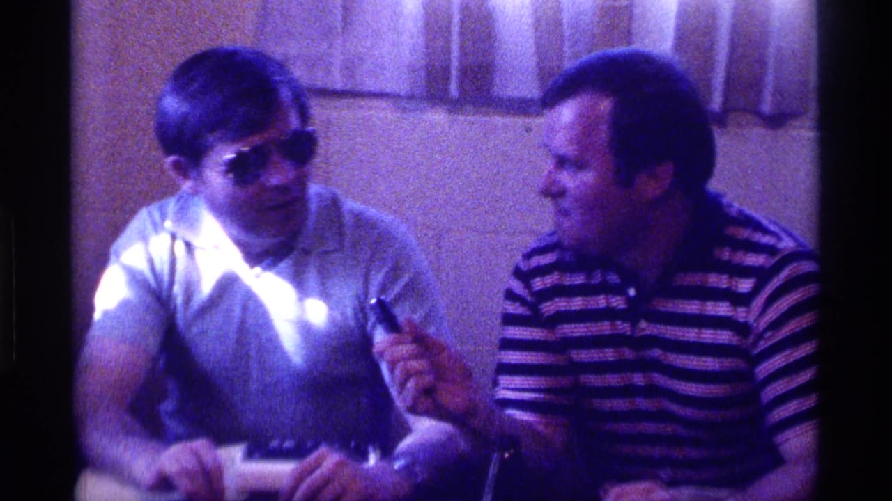 A man in sunglasses faces a man in a striped shirt holding a microphone.