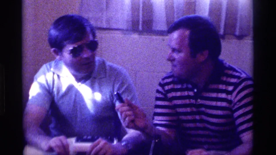 The man in sunglasses turns toward the interviewer as the microphone is held between them in a purple-tinted private room.