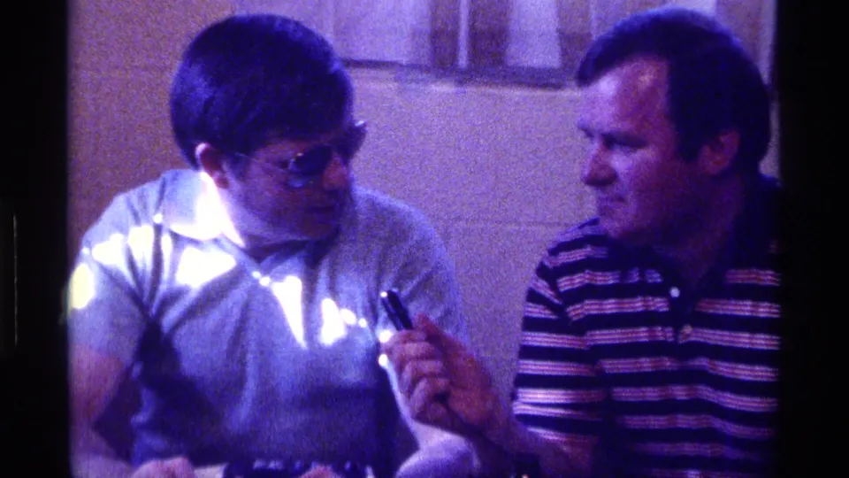 A close 8mm frame shows the sunglasses-wearing man looking down as the striped-shirt interviewer keeps the microphone raised.