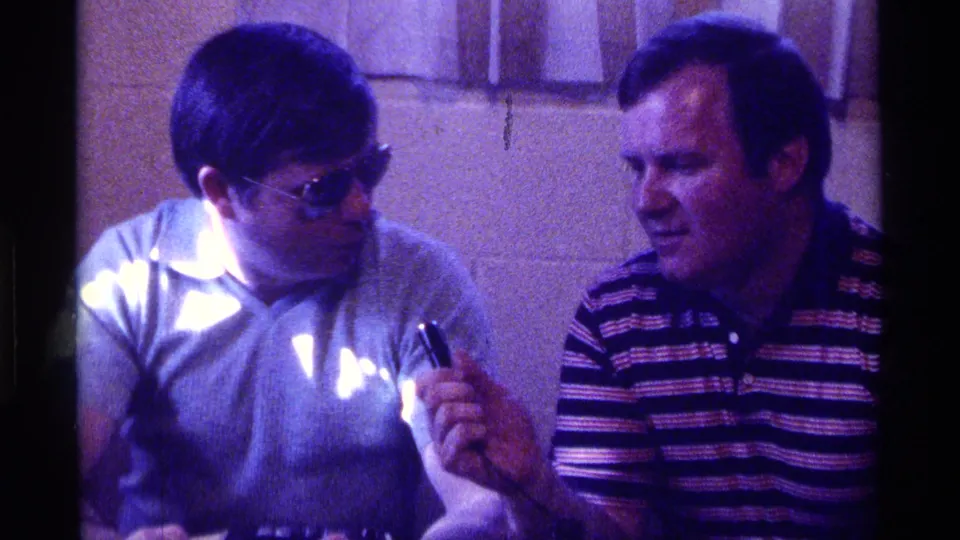 The two men appear to converse at the table, with the interviewer smiling slightly while holding the microphone in the dim room.