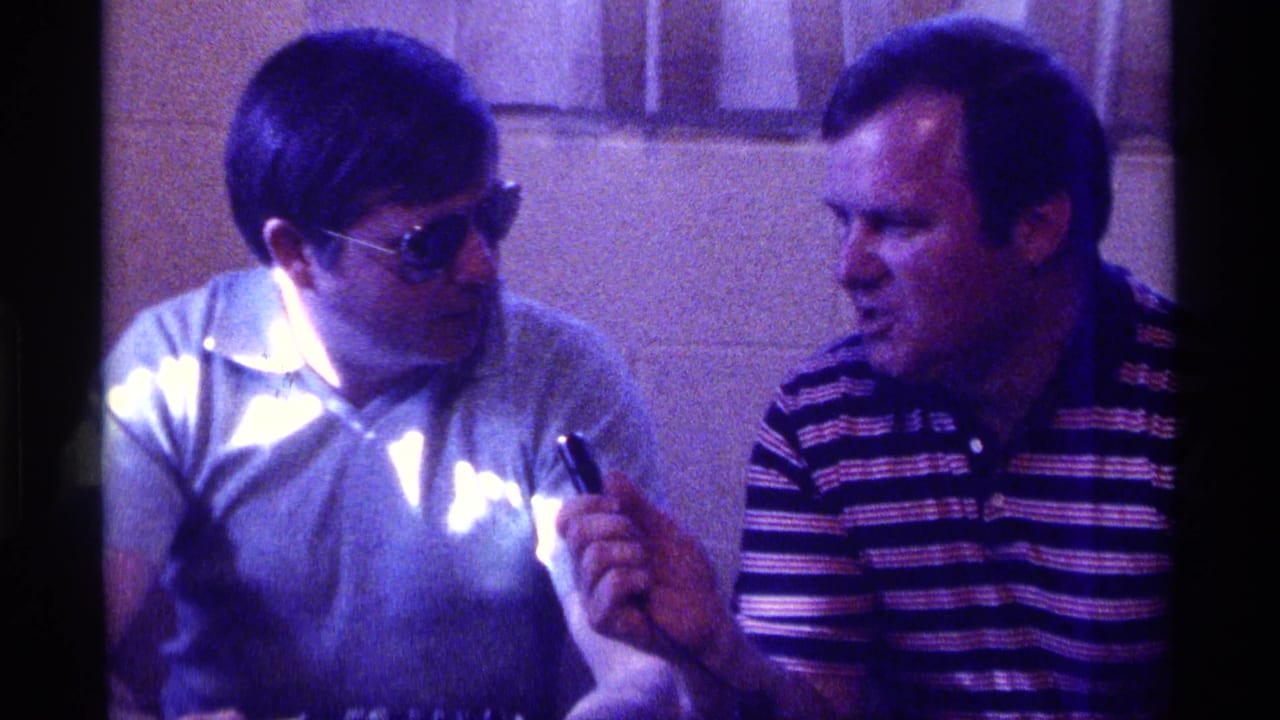 Two adult men sit indoors, one wearing sunglasses and holding a small object toward the other.