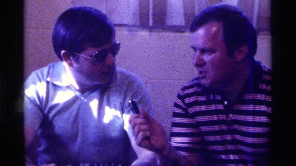 1976 Kentucky 8mm frame of two adult men in polo shirts conversing indoors, with one man in sunglasses holding a small object.